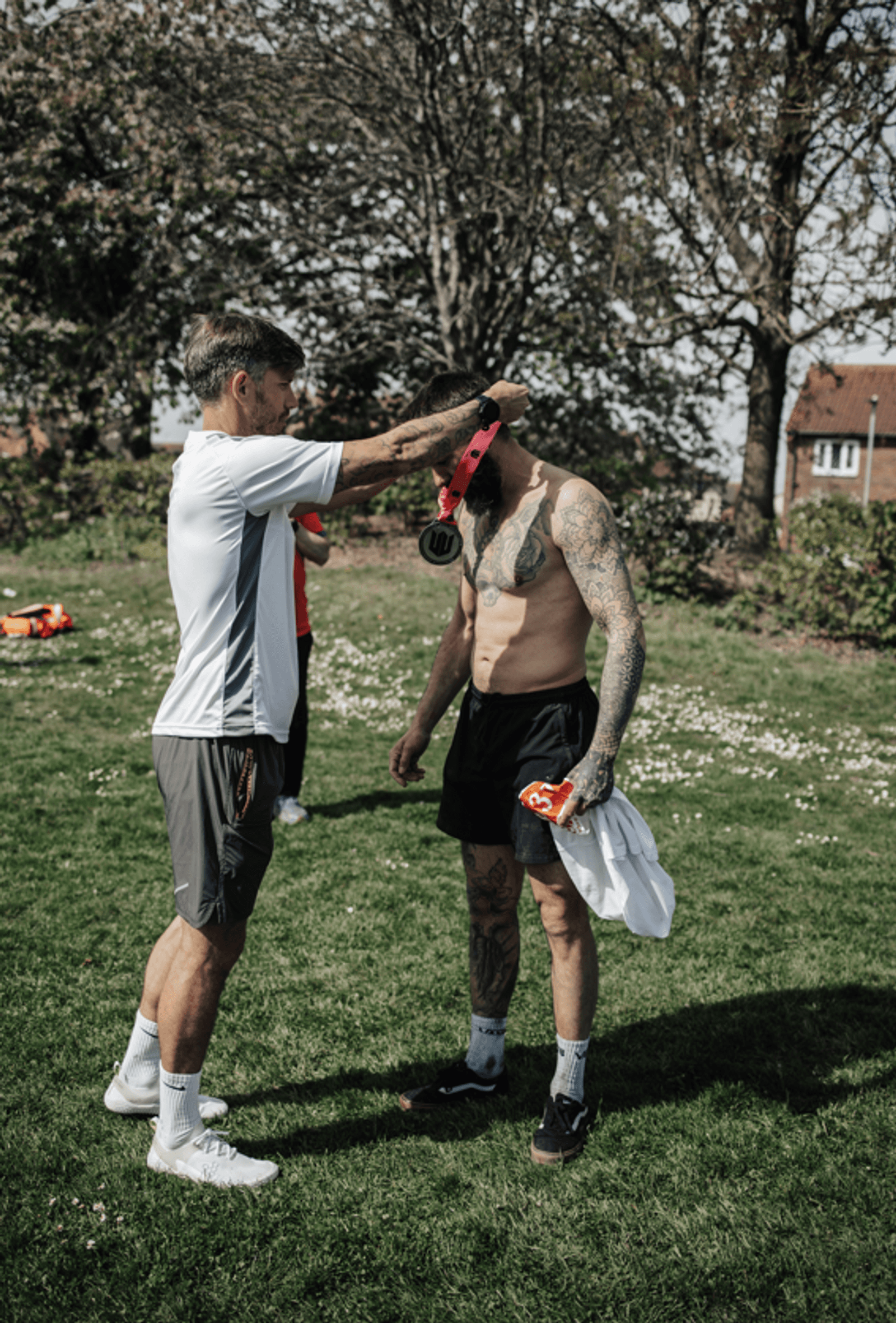 A man in a white shirt places a medal around the neck of a shirtless, tattooed man wearing black shorts and a cap, standing on grass outdoors with trees and buildings in the background.
