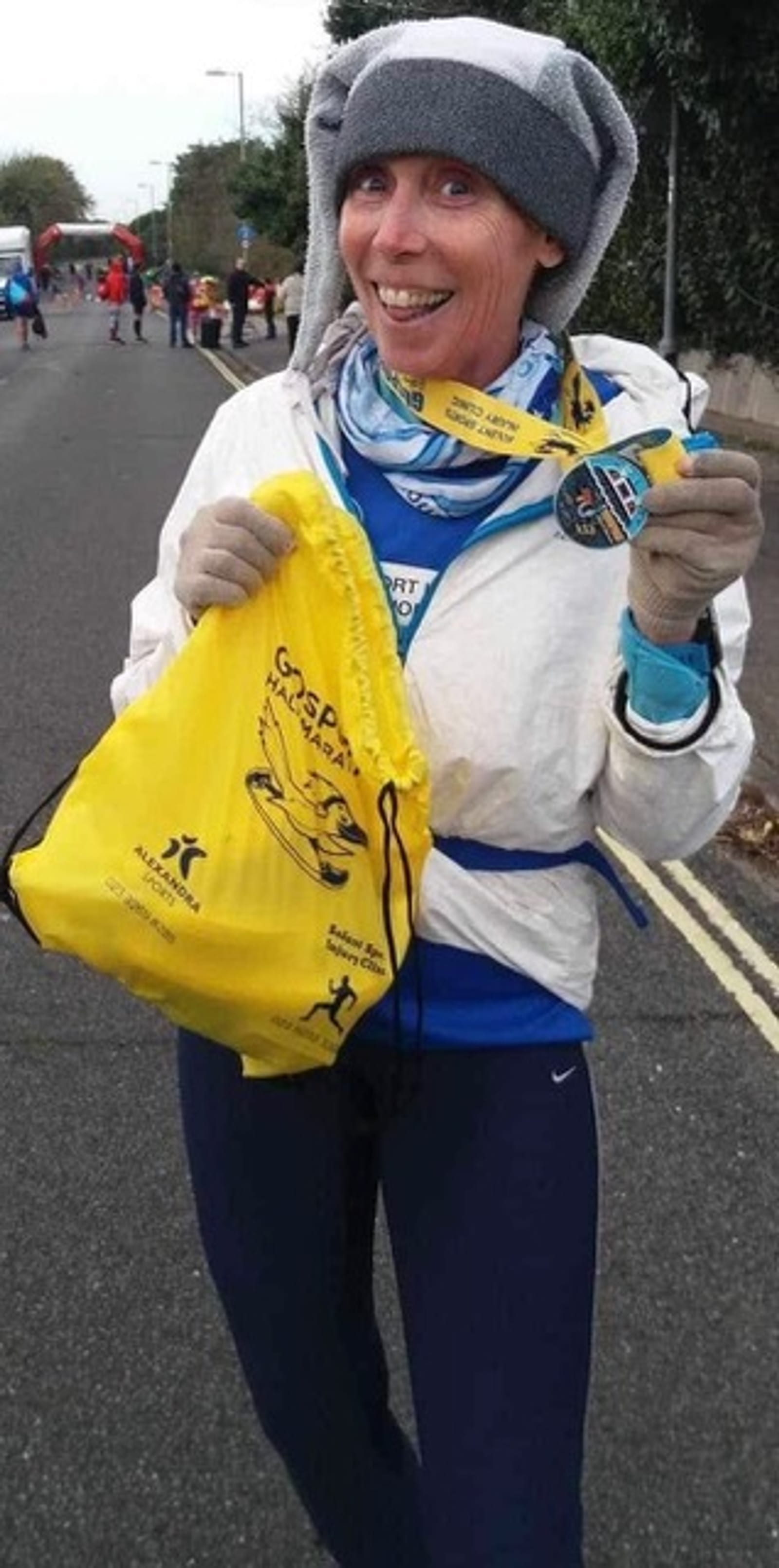 A smiling runner in a white jacket and blue hat holds up a medal and a yellow drawstring bag on a street after completing a race. Other participants and trees are visible in the background.