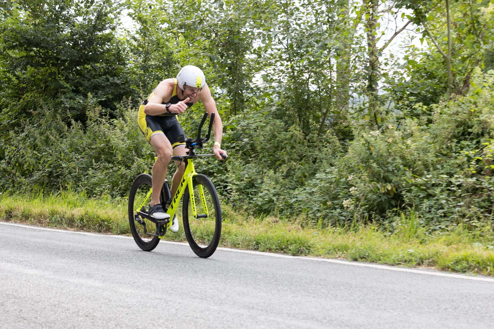 A cyclist wearing a yellow and black outfit and a white helmet rides a yellow bike on a road surrounded by green trees and bushes.