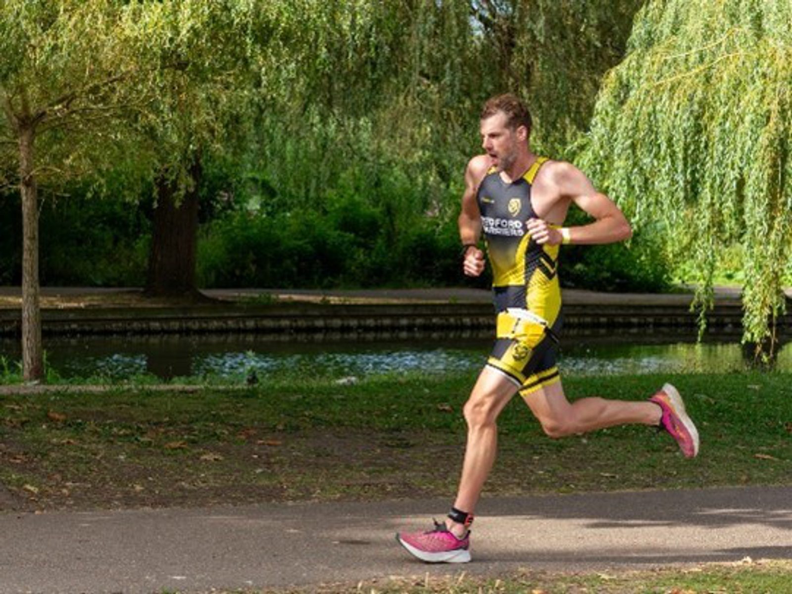 A male athlete in a yellow and black triathlon suit runs on a paved path beside a canal, with trees and greenery in the background on a sunny day.