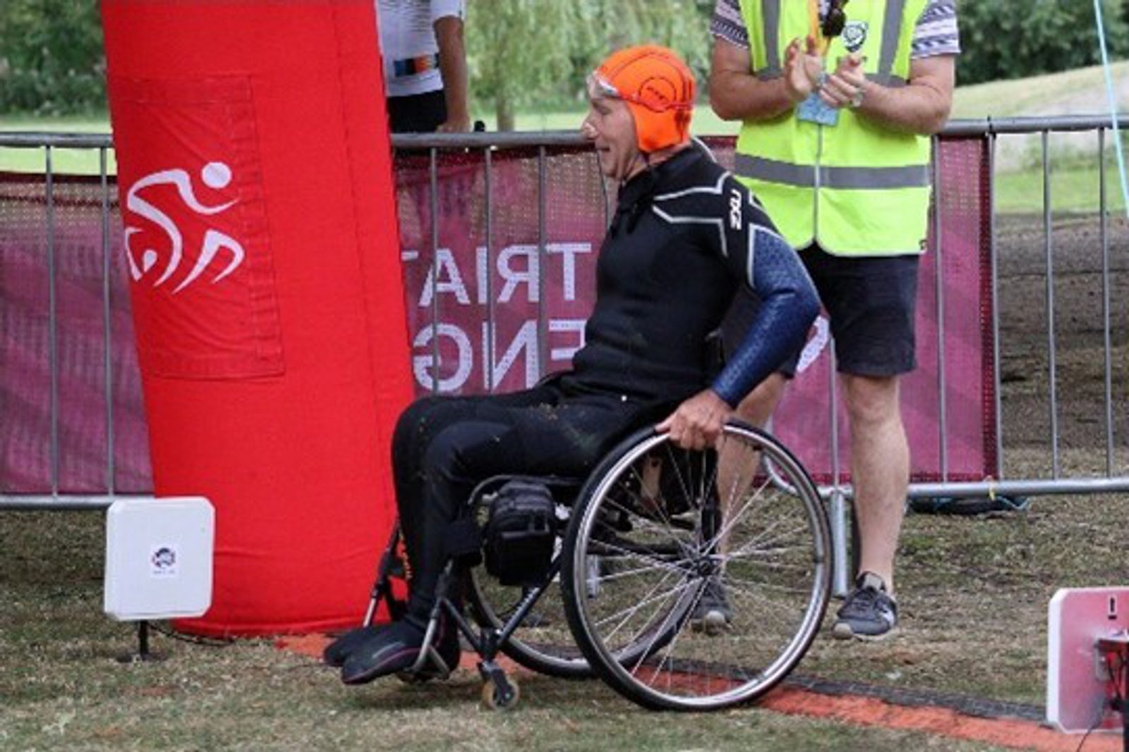 A triathlete in a wetsuit and orange swim cap sits in a wheelchair at the edge of a racecourse, near a red inflatable marker, with a volunteer standing nearby.