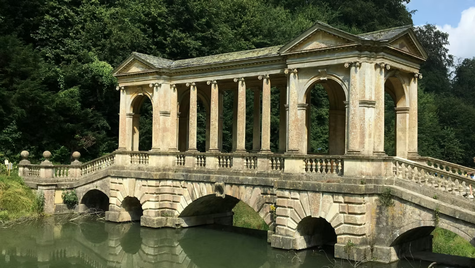A historic stone bridge with classical columns and arches crosses over a calm river, surrounded by lush green trees and vegetation. The bridge has a roofed section and decorative balustrades.