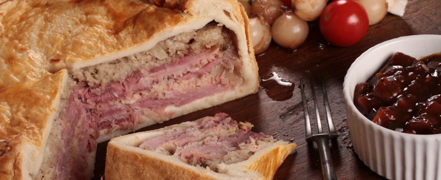 A sliced meat pie with layers of ham and stuffing on a wooden board, served with pickled onions, cherry tomatoes, and a ramekin of chutney. A fork rests nearby.