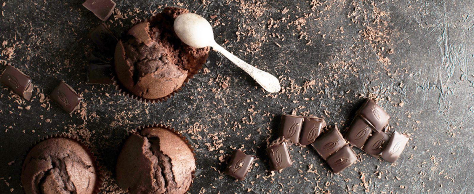 Two chocolate muffins and a small spoon on a dark surface, surrounded by pieces of chocolate and chocolate shavings.