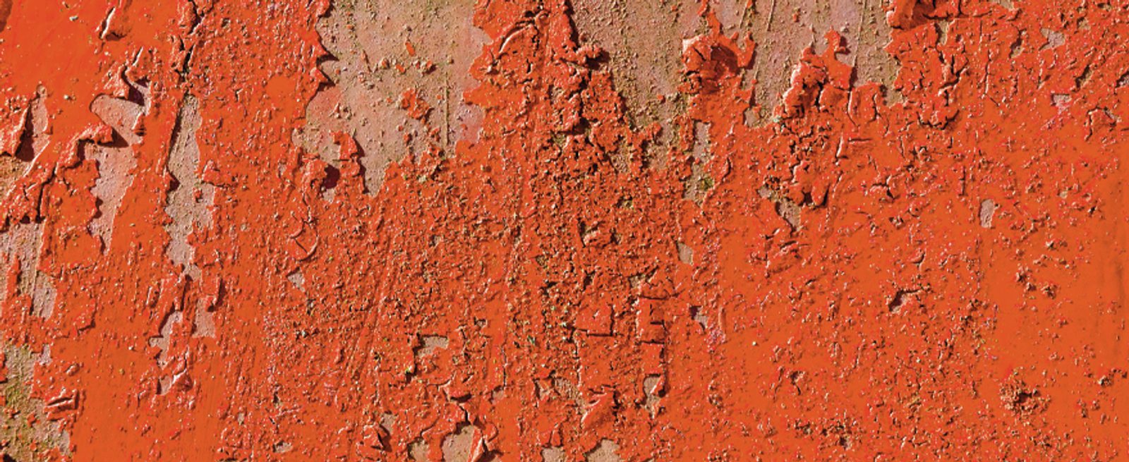 Close-up of a weathered surface with peeling and cracked red paint, revealing patches of exposed, rough brown material underneath. The texture appears uneven and flaking.