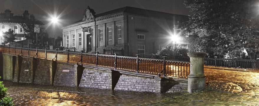 A nighttime scene of a historic brick building behind an old iron bridge. Streetlights cast a bright glow, highlighting the wet cobblestone pavement and creating a reflective effect. The image is mostly in black and white, except for golden hues on the bridge and pavement.