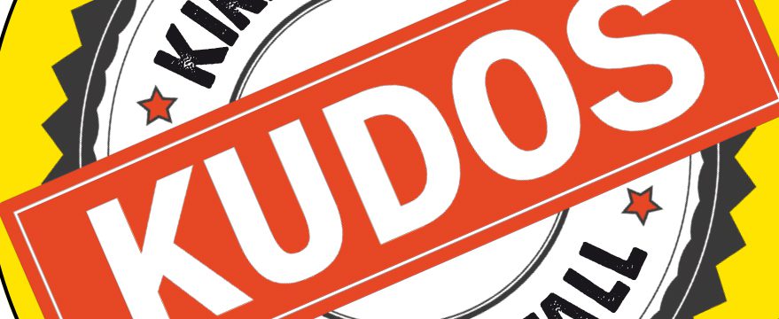 A circular badge with the word "KUDOS" in large white letters on a red banner across the center, with stars, and partially visible words "KIND" and "FULL" around the edges.