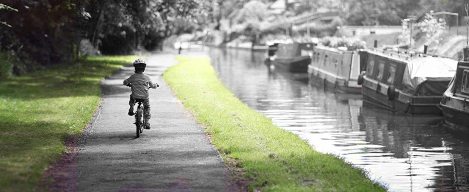 A child wearing a helmet rides a bicycle alone on a path beside a canal with boats moored along the water; the scene is mostly black and white except for the green grass.
