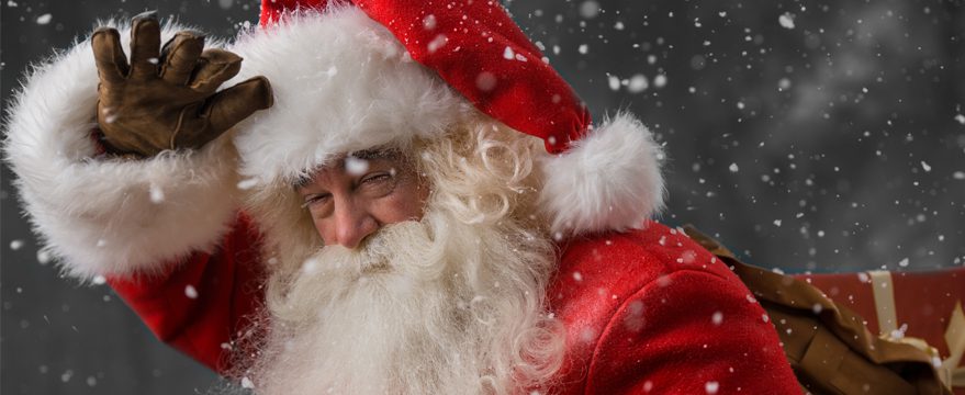 Santa Claus in a red suit and hat smiles and waves, with snow falling around him. He has a white beard and carries a brown sack over his shoulder against a dark background.