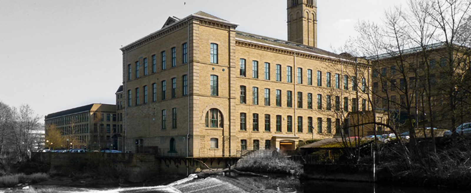 A large, historic stone mill building with tall windows and a prominent tower, situated by a river and surrounded by leafless trees and parked cars on a clear day.