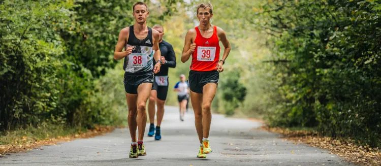 Two runners with race bibs 38 and 39 jog side by side on a tree-lined path, with other participants visible in the background during a race or marathon event.