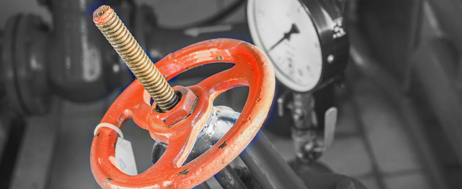 Close-up of a red industrial valve wheel with a threaded metal rod, in front of a blurred pressure gauge and pipes.