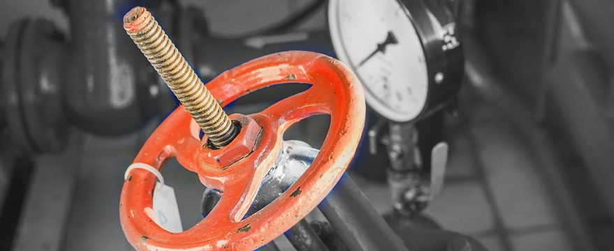 Close-up of a red industrial valve wheel with a threaded metal rod, in front of a blurred pressure gauge and pipes.
