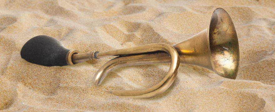 A brass horn with a black rubber bulb lies partially buried in sand, showing signs of wear and tarnish on its surface.