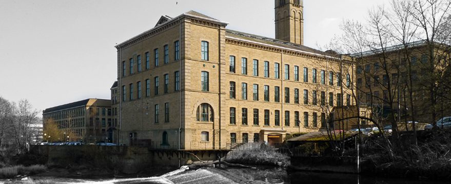 A large historic mill building made of light-colored bricks with many windows, located alongside a river. Leafless trees and parked cars are visible next to the building under a clear sky.