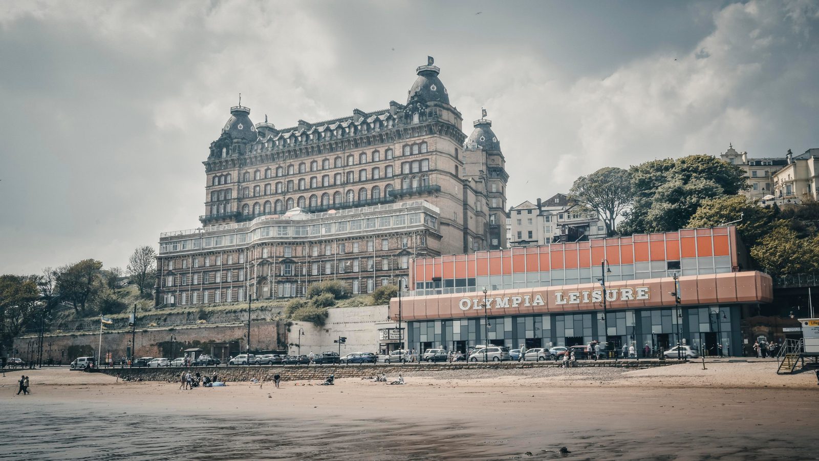 A large, historic hotel sits on a hill above a sandy beach, with the modern Olympia Leisure building in front. People walk and play on the beach under a cloudy sky, with trees and cars in the background.