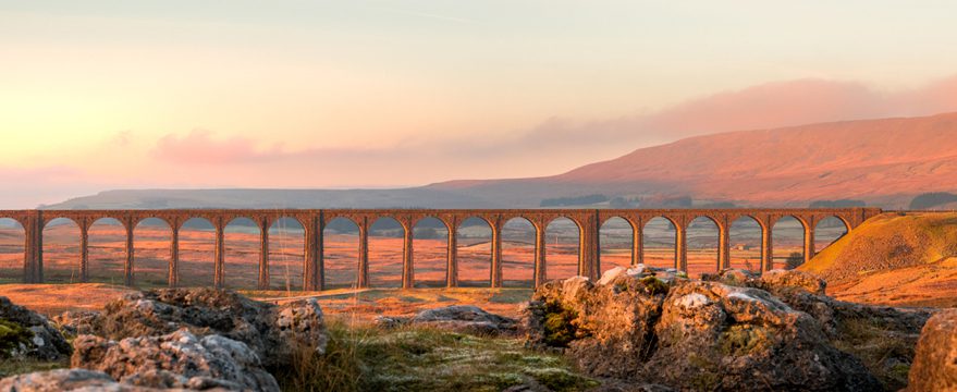 A long stone viaduct with multiple arches stretches across a sunlit, rocky landscape with rolling hills in the background under a soft, hazy sky.