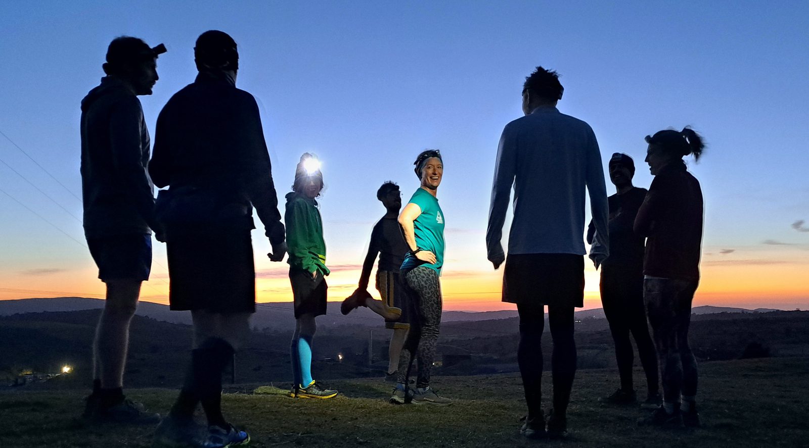 A group of runners stands on a hill at sunset, some silhouetted against the sky while one person in blue, illuminated and smiling, faces the camera. The landscape and fading sunlight create a scenic backdrop.
