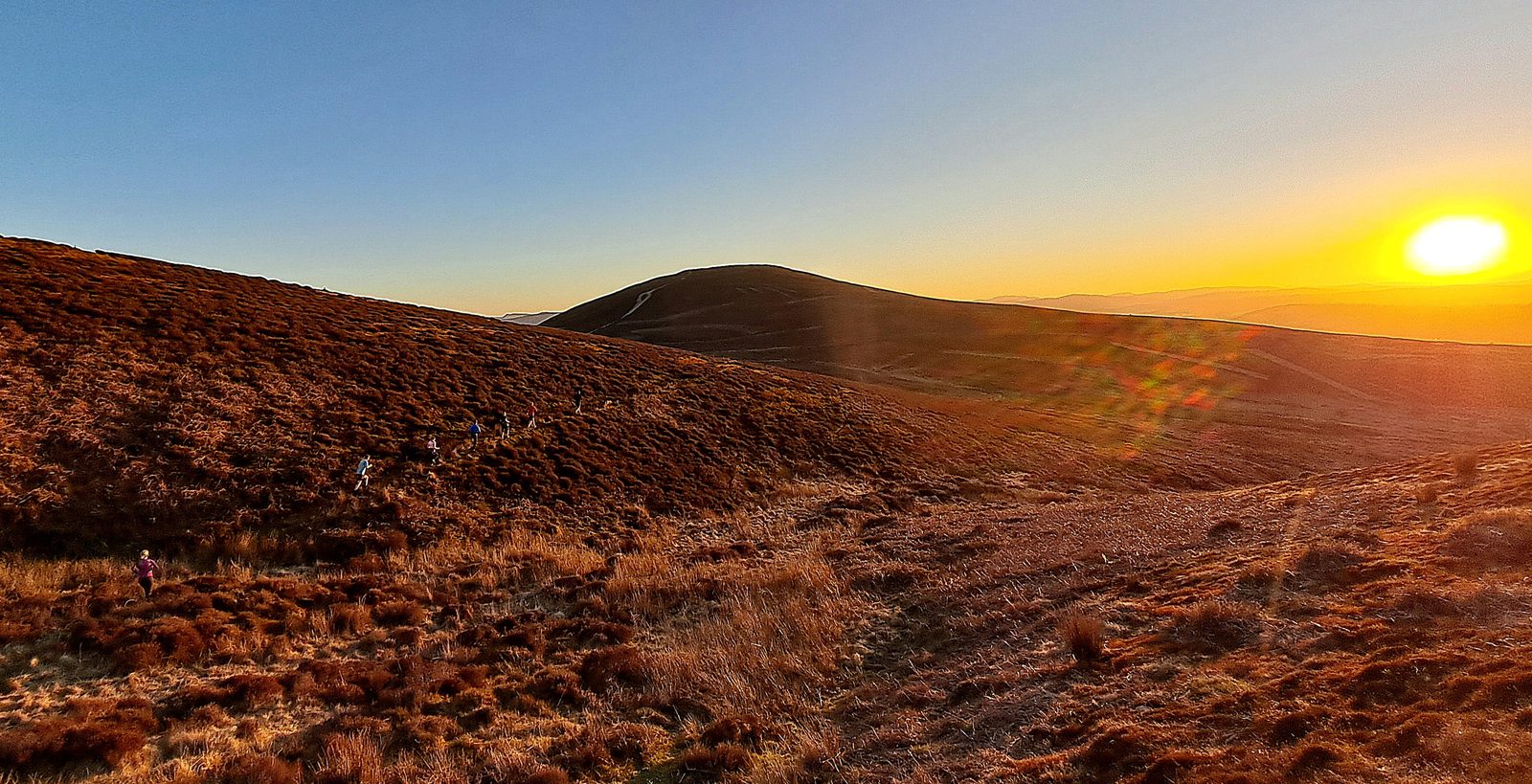 A group of hikers walk along a trail on a brown grassy hillside, with a mountain in the distance and the sun setting over the horizon, casting a golden glow across the landscape.