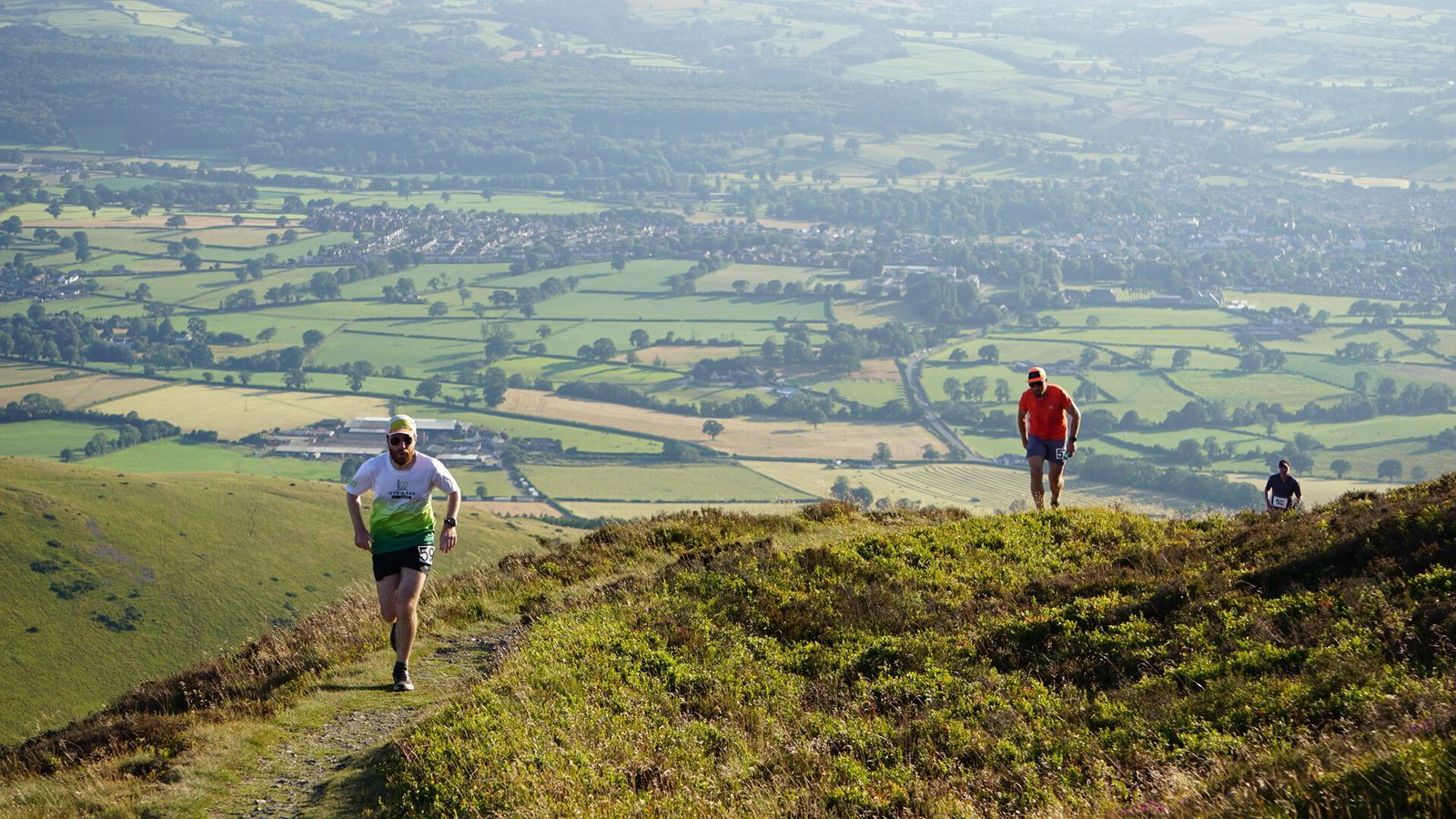 Three people hike up a grassy hill with a scenic countryside of fields, trees, and distant houses in the background under clear skies. The lead person is jogging while the others follow behind.