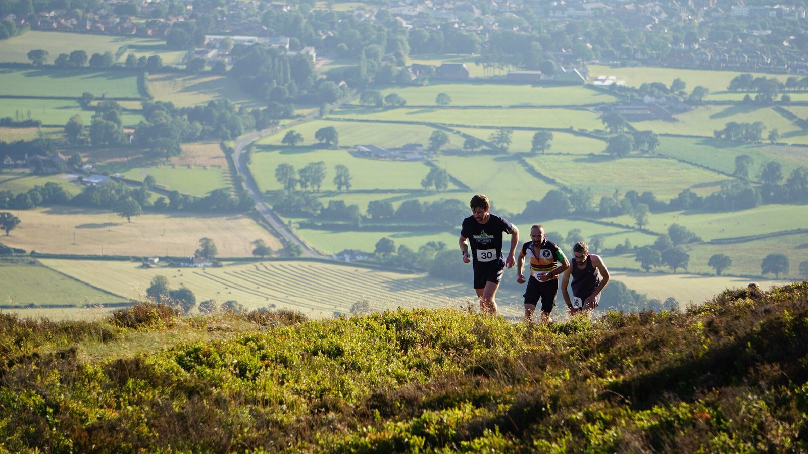 Four runners wearing numbered race bibs are climbing a grassy hill with a scenic, patchwork countryside landscape and village in the background under daylight.