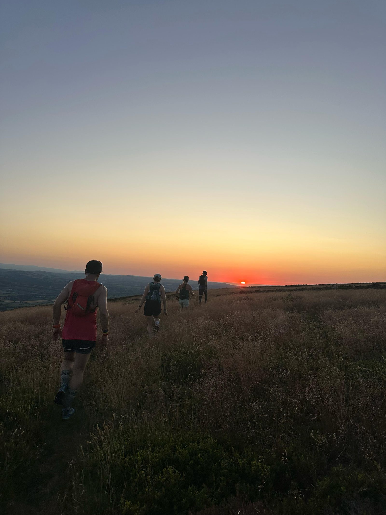 Four people hike along a grassy hill at sunset, with the sun low on the horizon and soft orange light illuminating the sky. The path winds through tall grass, and the landscape stretches out in the background.