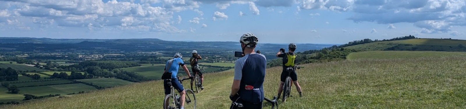 Four cyclists pause on a grassy hill overlooking a scenic valley with fields, forests, and distant hills under a partly cloudy blue sky. One cyclist takes a photo of the view.
