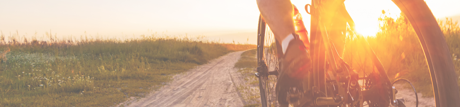 A close-up of a person riding a bicycle on a dirt path through grassy fields at sunset, with sunlight creating a warm, golden glow.