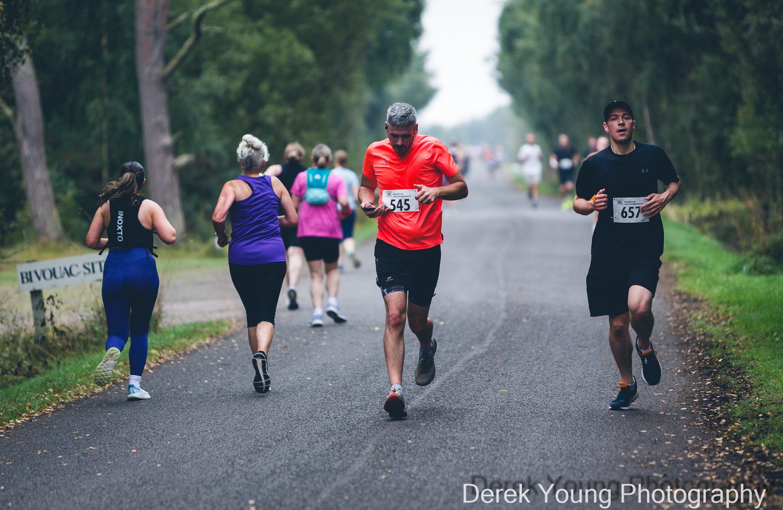 Runners participate in a race on a tree-lined road, wearing numbered bibs. Some are running while others jog at varying paces. The weather appears overcast, and greenery surrounds the path.