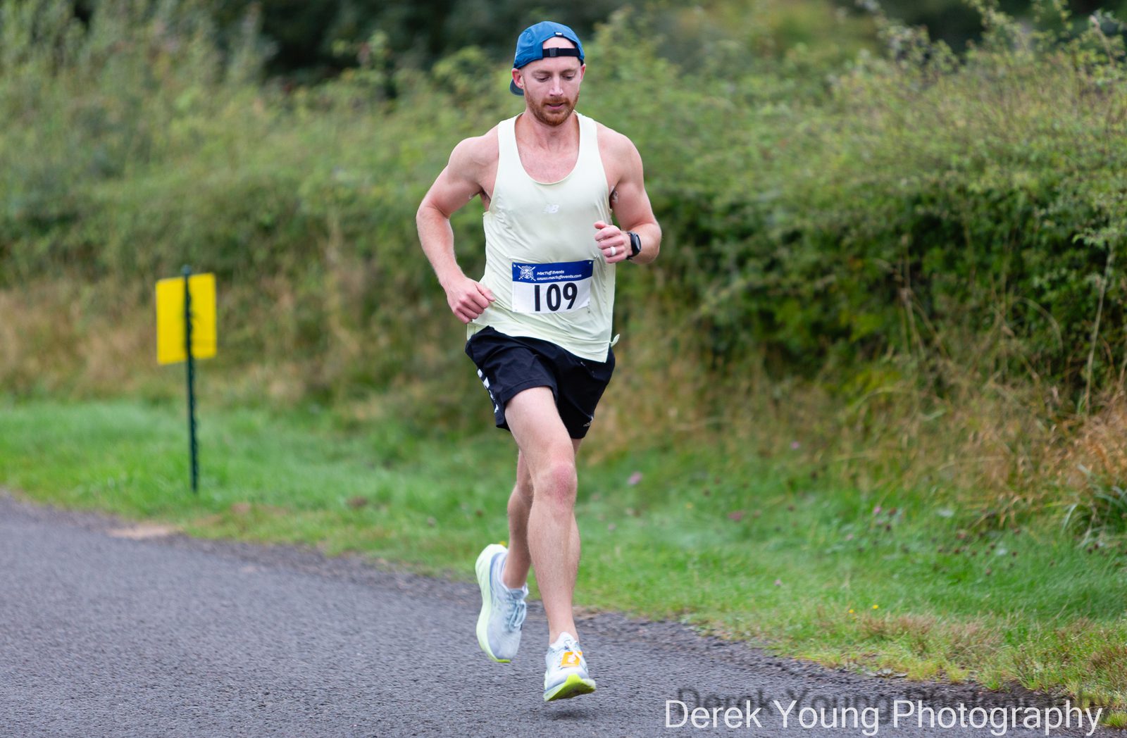 A male runner wearing a white tank top, black shorts, and a blue cap runs on a paved path beside greenery. He is wearing bib number 109. “Derek Young Photography” is visible at the bottom right.