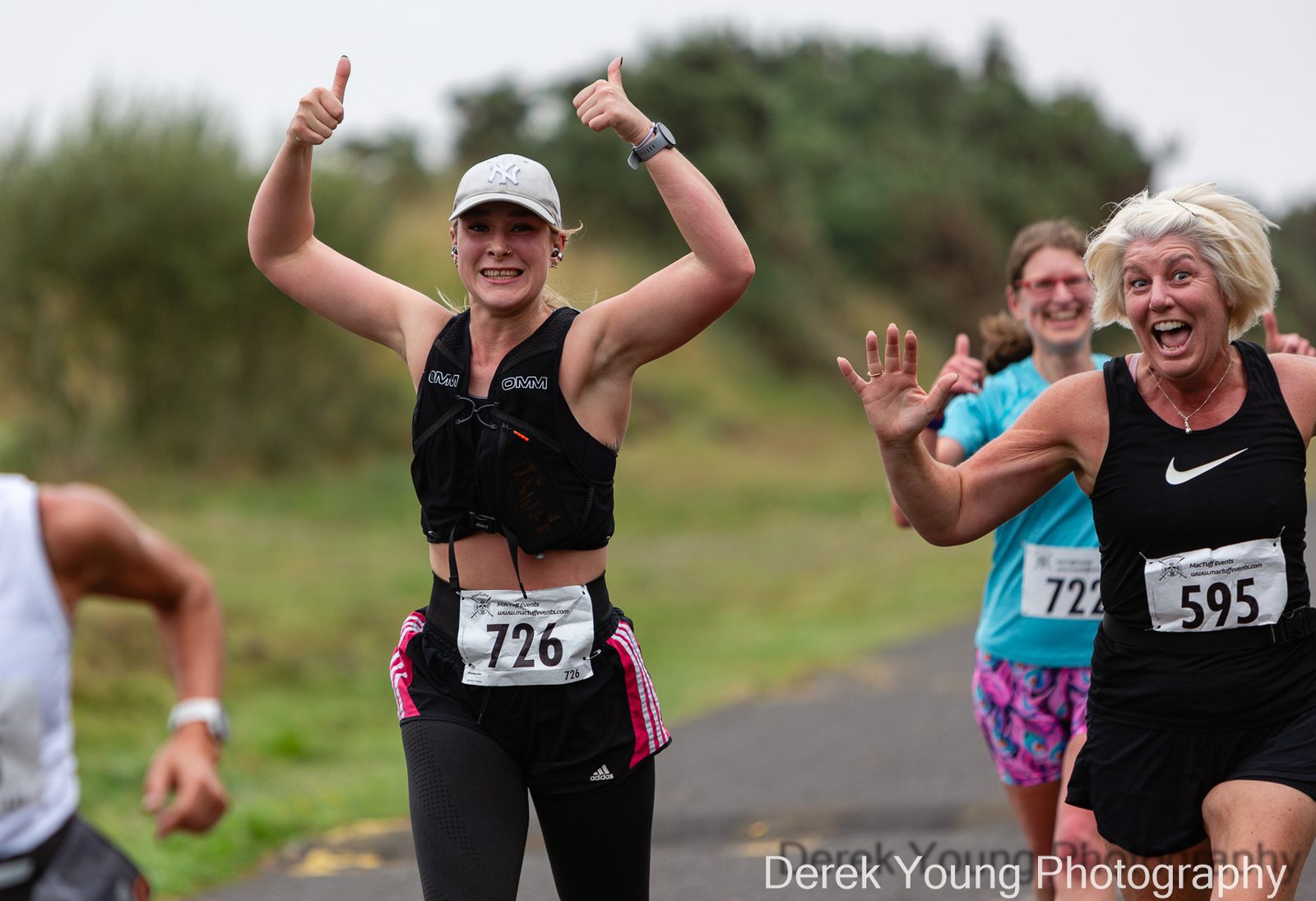Three women are running a race outdoors, smiling and giving thumbs up toward the camera. They wear athletic clothing and race bibs, with greenery in the background. The atmosphere appears energetic and joyful.