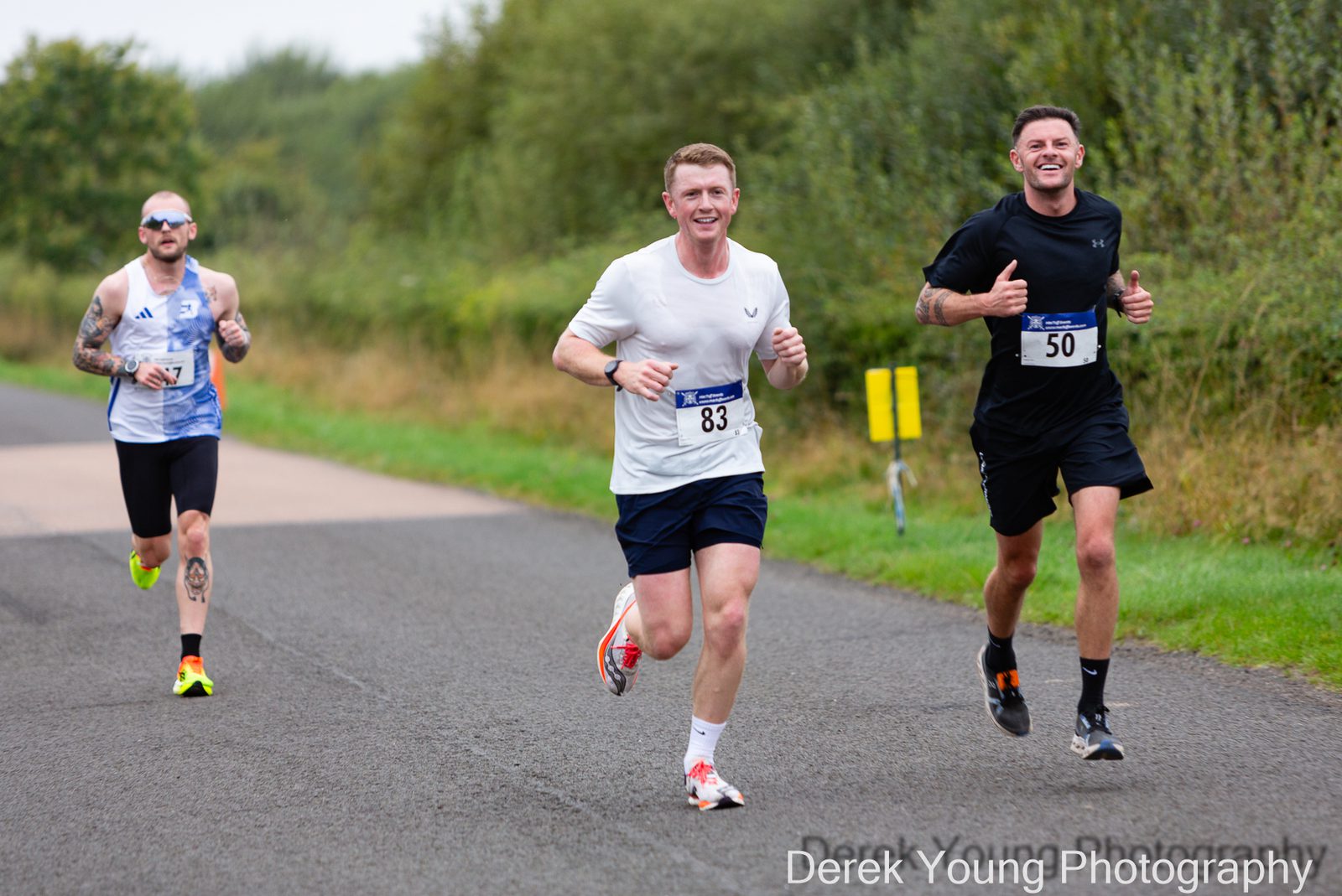 Three male runners participate in a road race, smiling and giving thumbs up as they run on a paved path surrounded by greenery. Two runners wear numbered bibs, and one has tattoos and sunglasses.