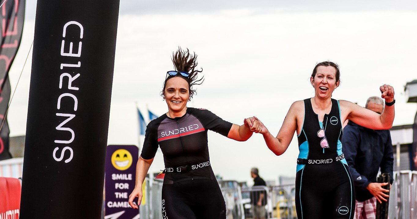 Two women in wetsuits cross a finish line holding hands, smiling and celebrating. A black banner with "Sundried" in white text and a sign with a smiley face are visible in the background.