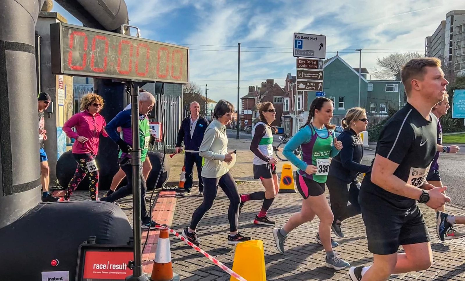 A group of people are running in a race on a sunny day. Some are near the starting line marked by a timer that reads "00:00:00." Participants wear various athletic clothing, each with race numbers. In the background are buildings, signs, and onlookers.