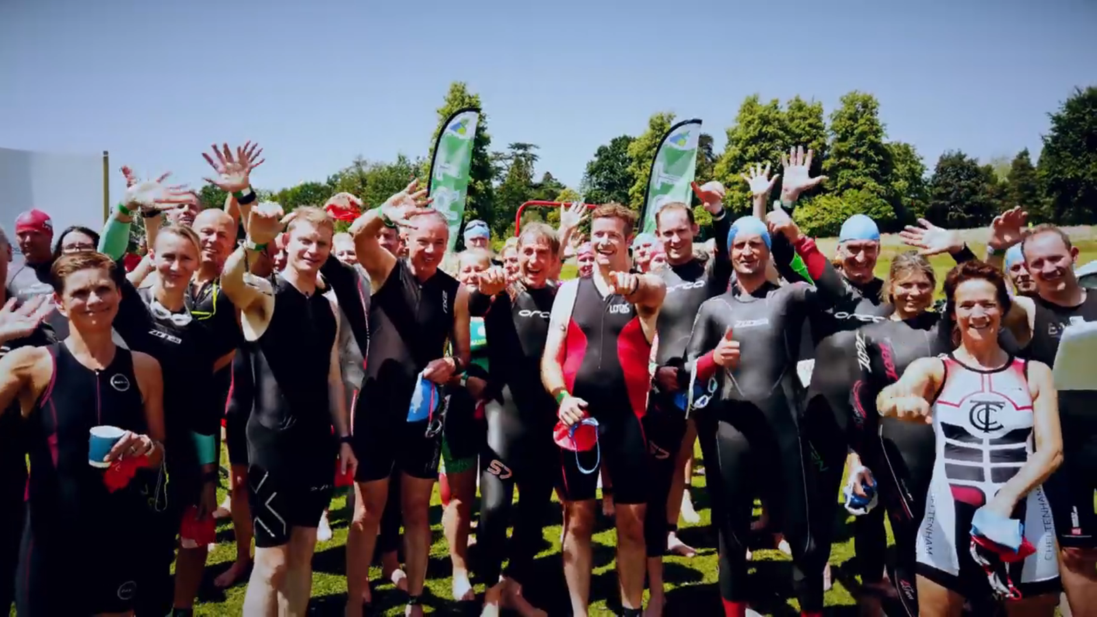 A group of triathletes stands outdoors on a sunny day, smiling and waving at the camera. They are wearing wetsuits and holding swim caps. Behind them, there are green and white flags and trees. Everyone looks enthusiastic and ready for the event.