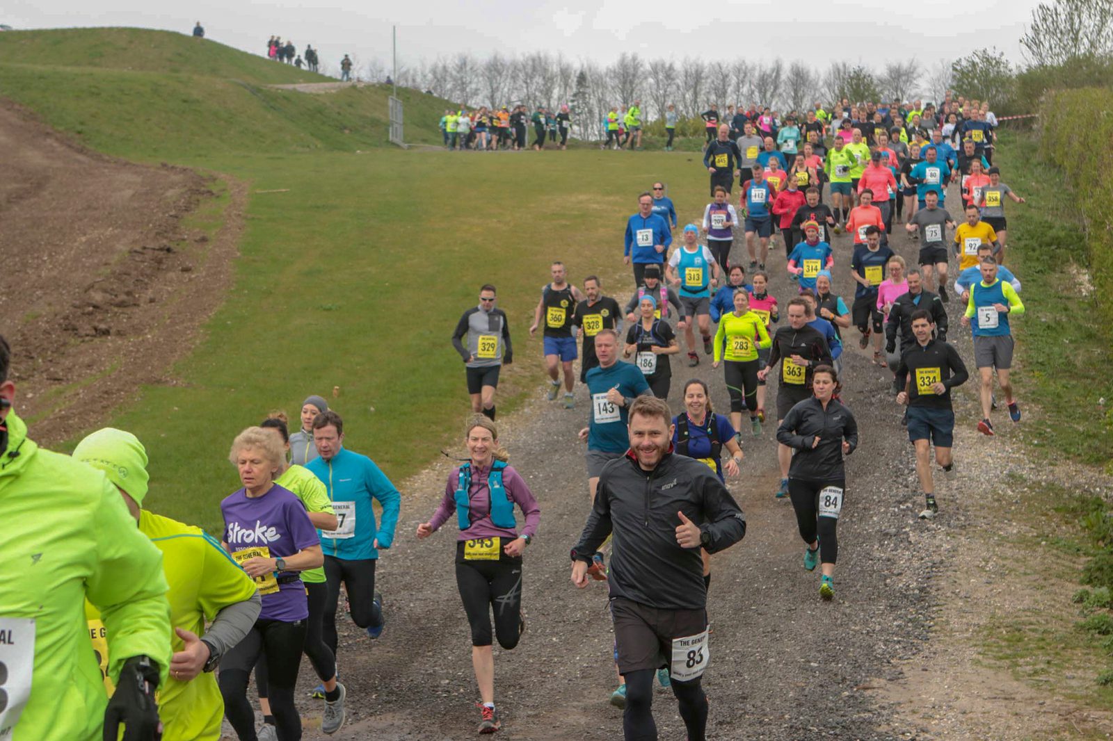 A large group of runners participating in a race on a trail with grass and sparse trees in the background. They wear various colored outfits and bib numbers, some giving thumbs up. The sky is overcast, and there are more people in the distance behind them.
