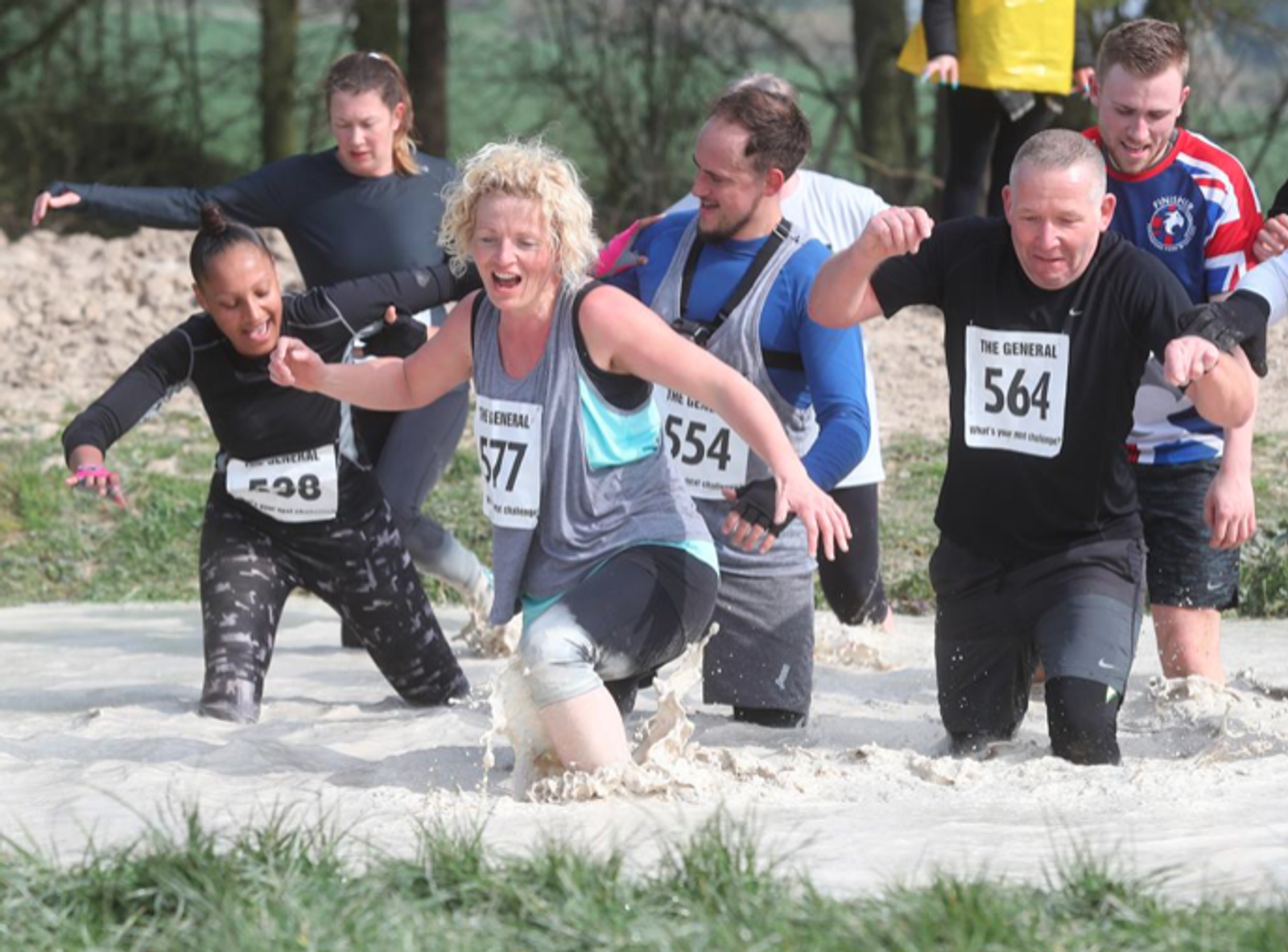 A group of people wearing athletic clothing and race numbers wading through a muddy water obstacle during an outdoor challenge race. They appear to be having fun and are exerting effort to move through the muddy terrain. Trees and grass are visible in the background.