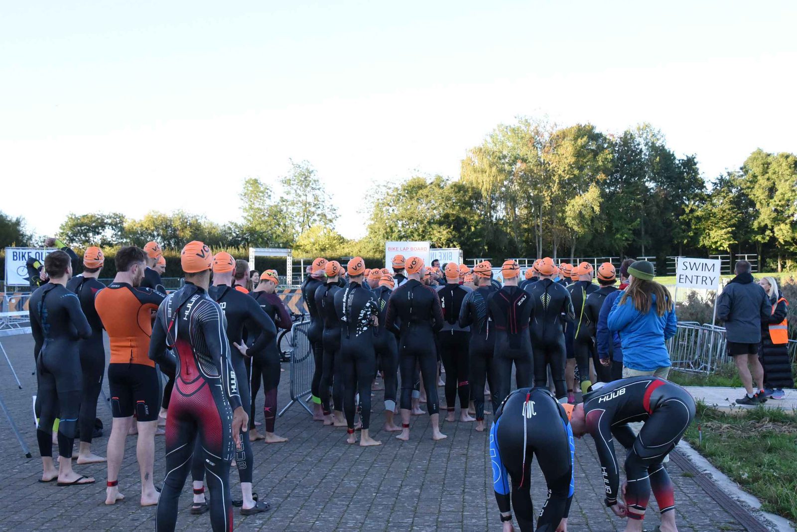 A group of athletes in wetsuits and orange swim caps stand together outdoors near signs for “Swim Entry” and “Bike In,” preparing for a race, with trees and barriers in the background.