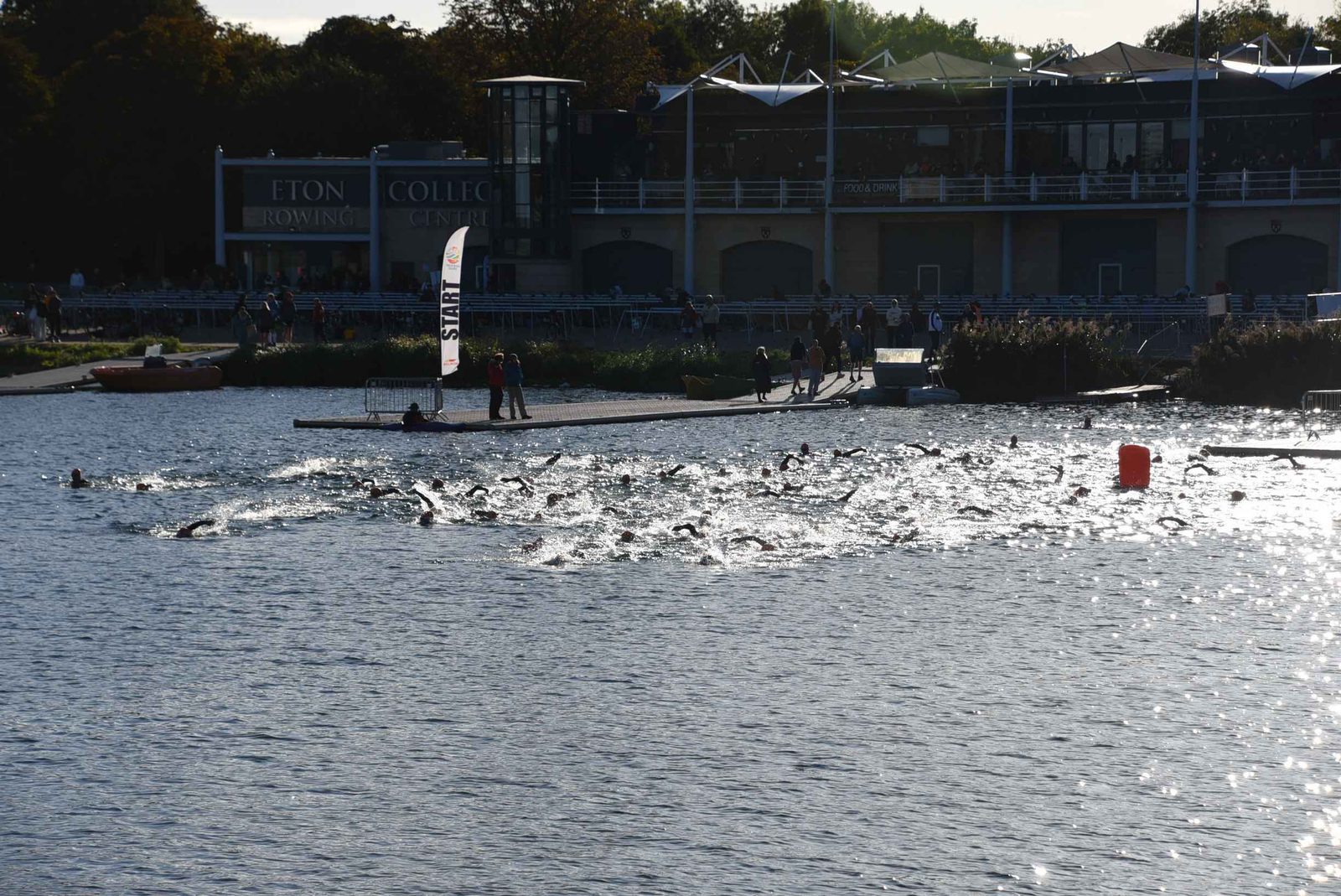 A large group of swimmers participates in an open water race near a dock, with spectators and officials watching from the shore in front of a building labeled "Eton Rowing." The water is splashing as swimmers move forward.