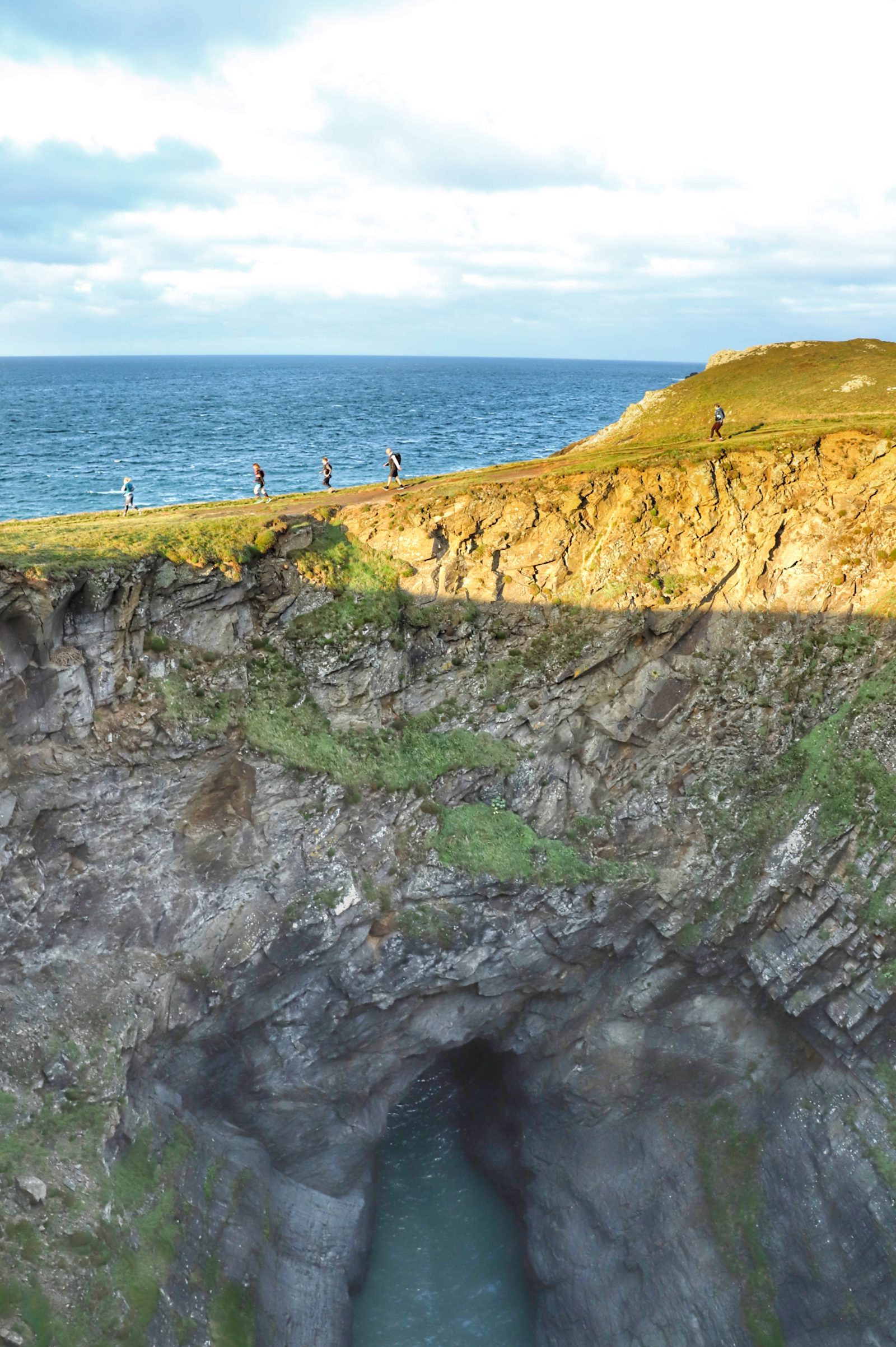 Five people walk along the edge of a cliff with a large rock formation and a sea cave below, with the ocean and a partly cloudy sky in the background. The sun casts a strong shadow across the cliff face and the rocky terrain.