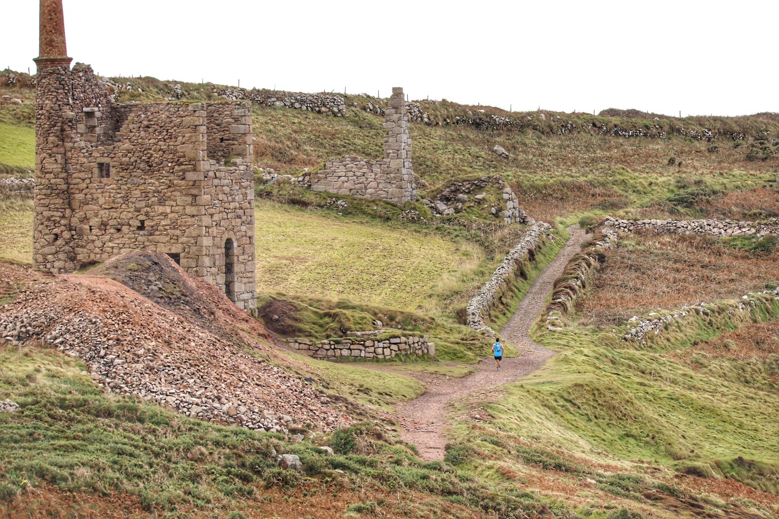 A lone person wearing a blue jacket walks on a dirt path beside the ruins of an old stone building with a tall chimney. The scenic landscape has patches of green grass, brown bracken, and stone walls running along the rolling hills under an overcast sky.