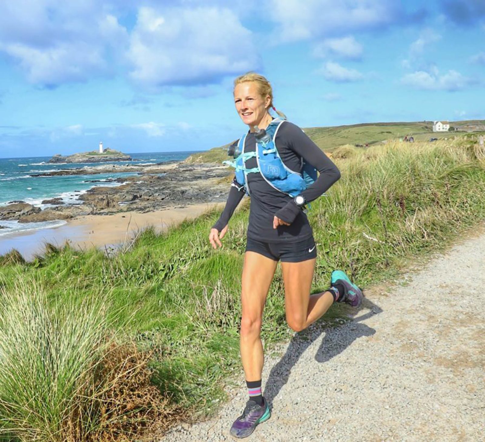 A woman is running along a gravel path near a scenic coastal area. She is wearing a black long-sleeve top, black shorts, a blue hydration pack, and running shoes. The background features grassy dunes, a rocky shoreline, and a lighthouse in the distance. The sky is partly cloudy.