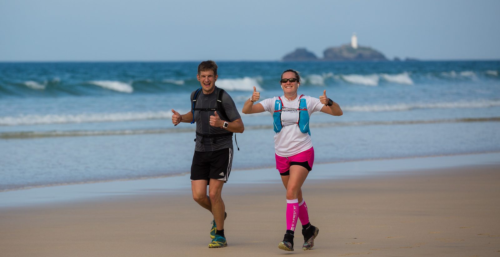 Two people are running on a sandy beach beside the ocean, both smiling and giving thumbs up. The person on the left is wearing a black outfit, and the person on the right is wearing a white shirt, pink shorts, and pink leg sleeves. An island with a lighthouse is visible in the background.