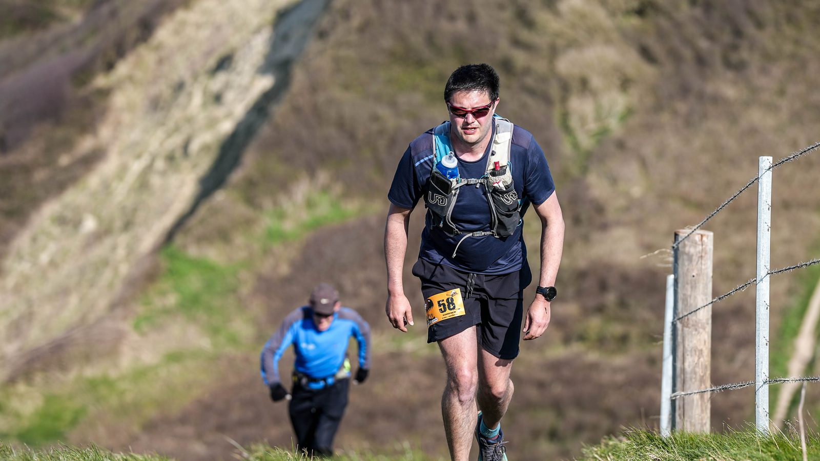 Two runners participating in an outdoor race traverse a grassy hillside. The forefront runner wears a dark athletic outfit, backpack, and race bib numbered 518, while the second runner, dressed in blue, follows in the background. Sunlit hills and a fence are visible.