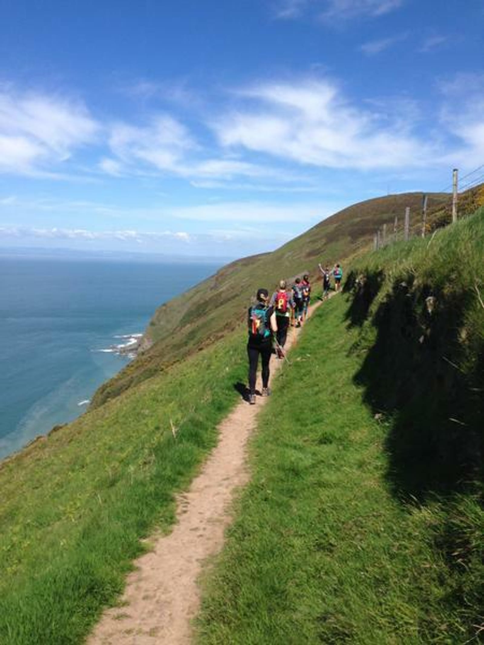 A group of hikers walks along a narrow dirt trail on a grassy hillside overlooking the ocean. The sky is clear with scattered white clouds. The hikers carry backpacks and are spaced out along the path, heading towards the horizon.