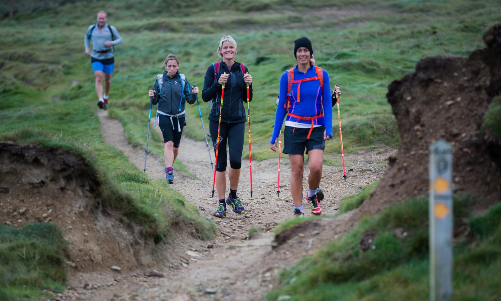 Four people hike on a trail through grassy terrain. Two women in the front walk with trekking poles and wear jackets and shorts. Another woman and a man follow behind, also with trekking poles, dressed in jackets and shorts. The weather appears cool and overcast.