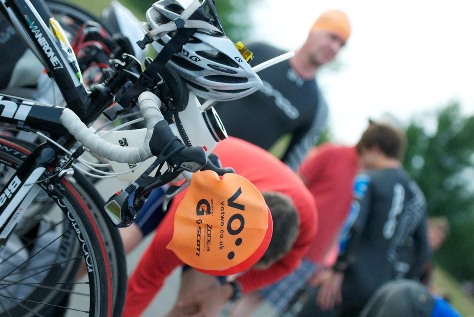 Close-up of a bicycle with a bright orange swim cap hanging from the handlebars. Blurred in the background, athletes prepare for a triathlon, some wearing wetsuits and swim caps. A green, outdoor environment suggests they are at the starting point.