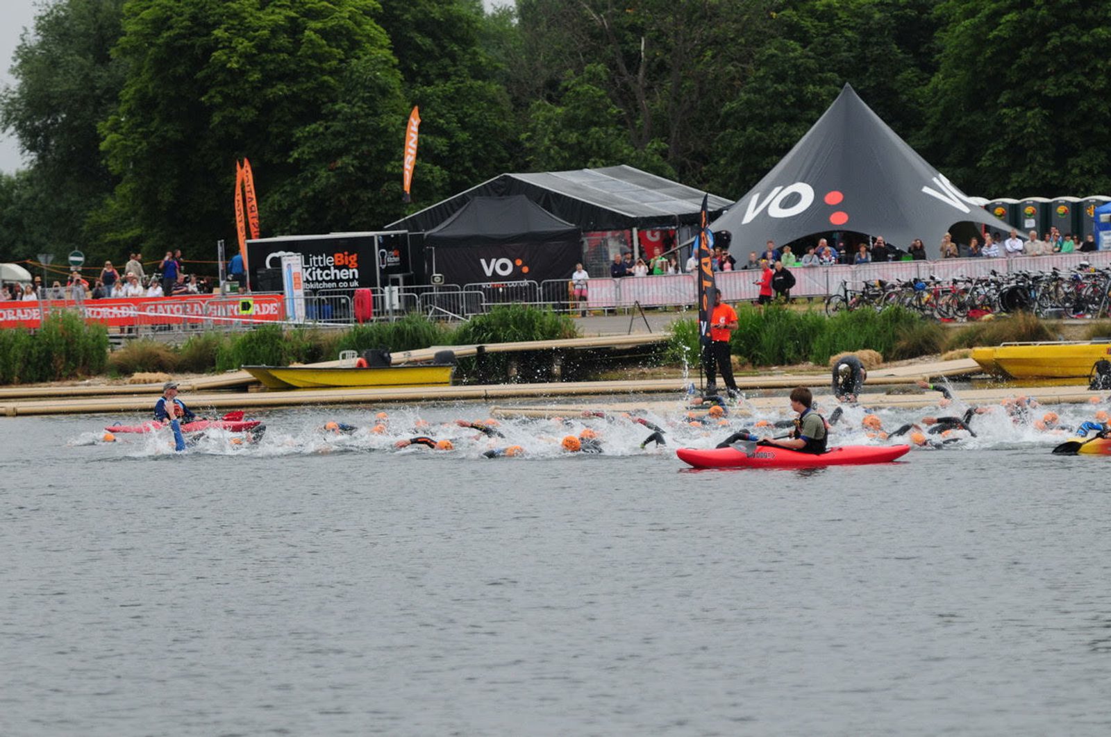 A group of swimmers competes in a triathlon, swimming towards the shore in a lake. Kayakers and rescue personnel observe from the water. A large tent branded with "VO:" and spectators are visible in the background amidst greenery.
