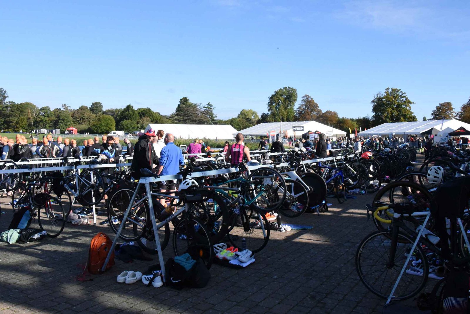 Dozens of bicycles are racked in a transition area at an outdoor sporting event, with athletes preparing and changing gear. Tents and spectators are visible in the background under a clear blue sky.
