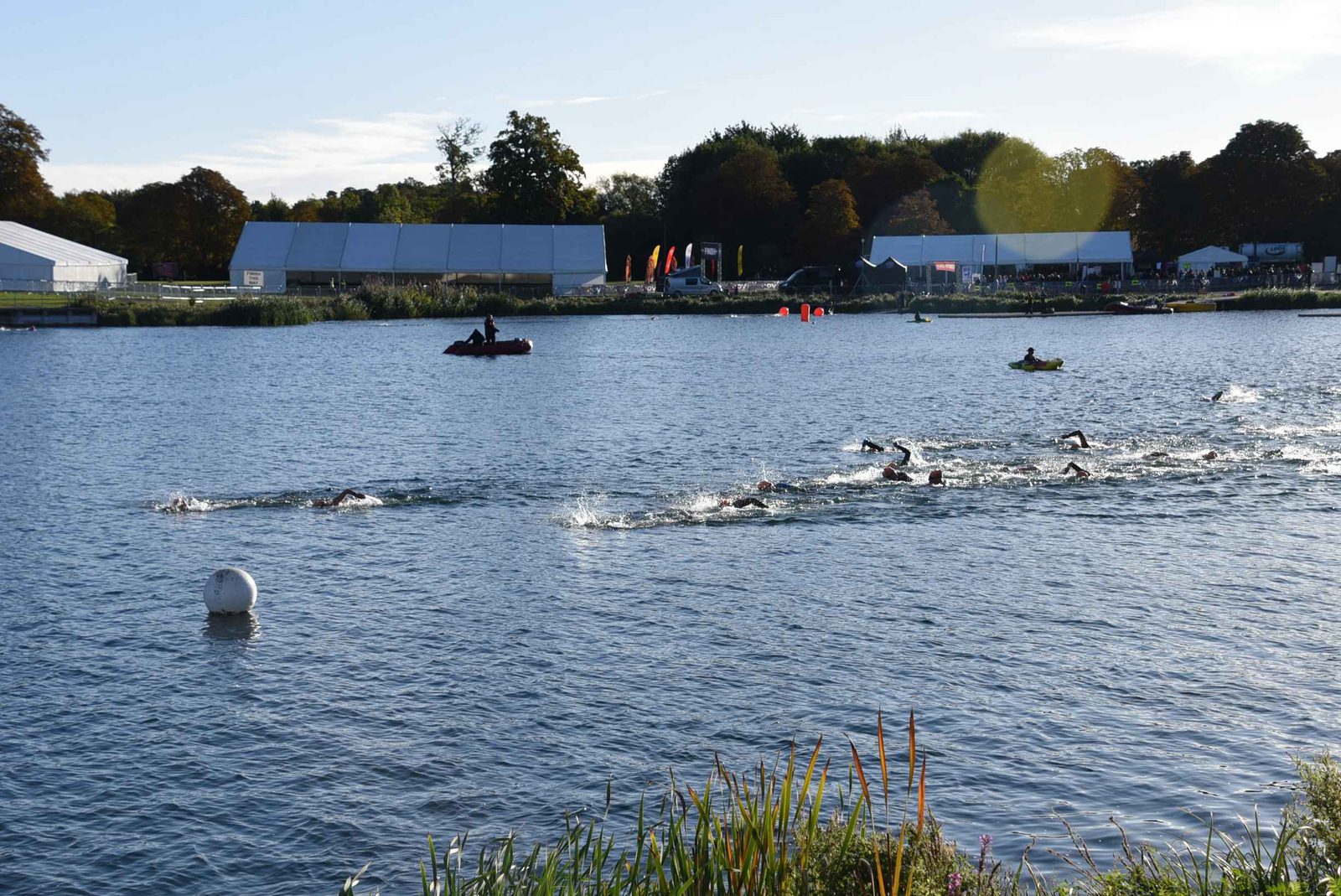 A group of swimmers compete in an open water race on a lake, with safety boats nearby. Event tents and trees line the far shore under a partly cloudy sky.